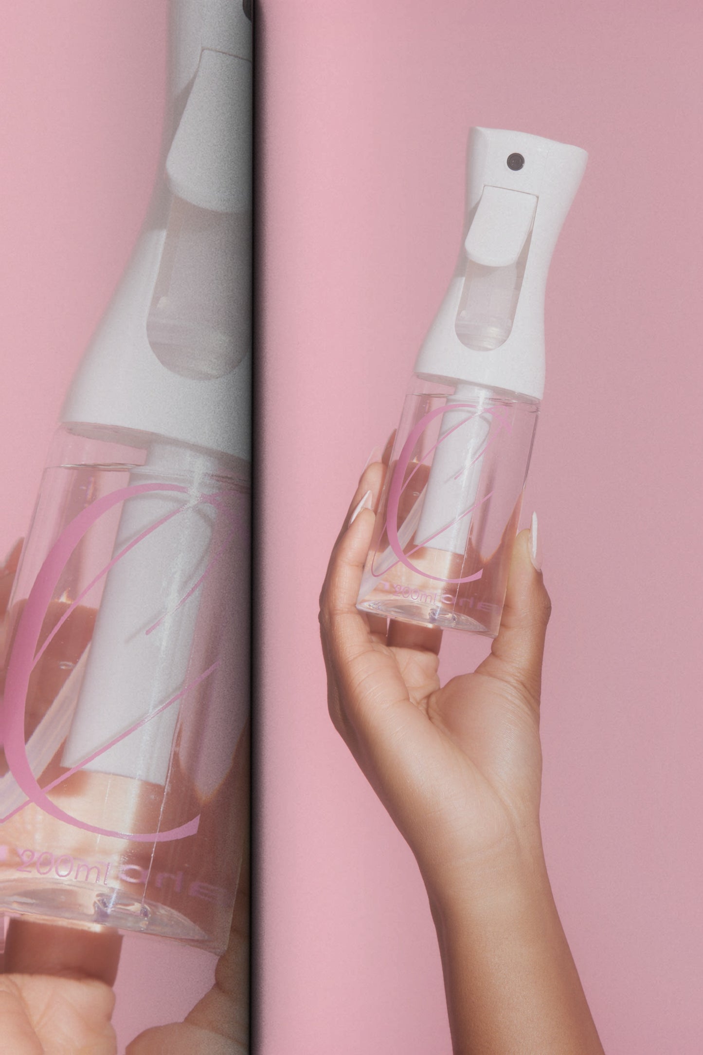Hand holding a clear spray bottle with pink accents against a pink background